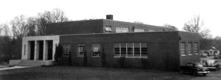 Black and white photograph of Oak Street Elementary School taken in 1954 for the Fairfax County School Board's fire insurance survey of school properties. The building has two stories in an 'L' shape. The main entrance is made of white stone and visually looks very different from the brick exterior. A pair of 1950s era cars are visible on the right side of the image. 