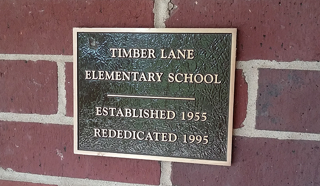 Photograph of the rededication plaque attached to the brick wall at the main entrance to our school. It reads Timber Lane Elementary School, Established 1955, Rededicated 1995.