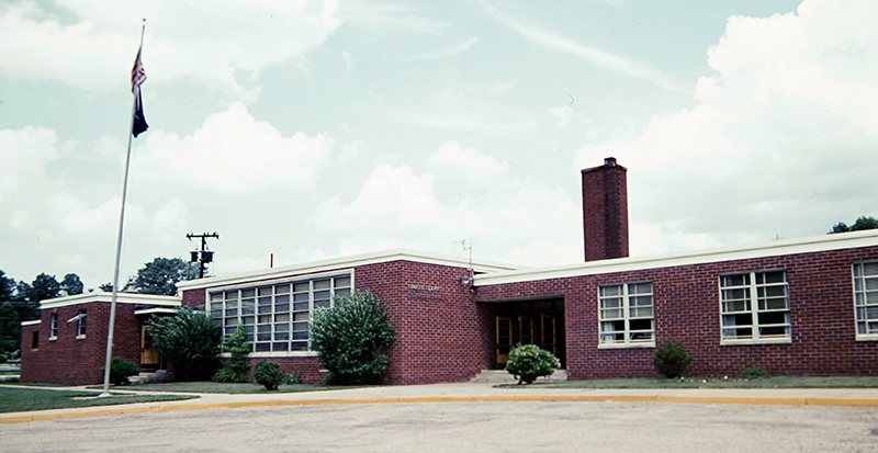 Color photograph of the old main entrance to Timber Lane Elementary School taken in the late 1970s or early 1980s. This section of the building looks much as it does today. The plants and shrubs pictured in front of the building have since been removed. The flag pole, on the left side of the picture, has since been moved to the new main entrance on the south side of the building. 